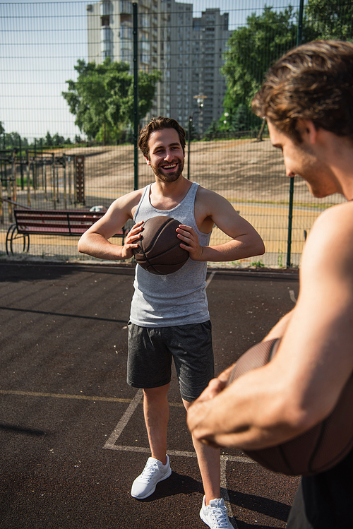 Smiling man holding basketball ball near blurred friend on court