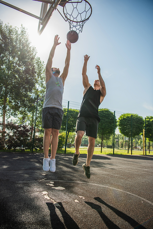 Young men jumping near ball and basketball hoop outdoors