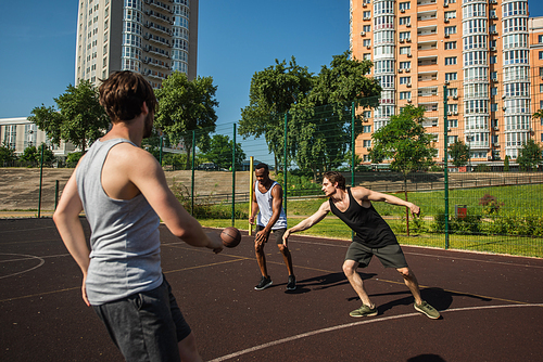 Interracial men playing basketball near blurred friend on playground