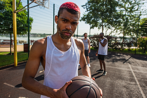 African american basketball player holding ball near blurred men on playground