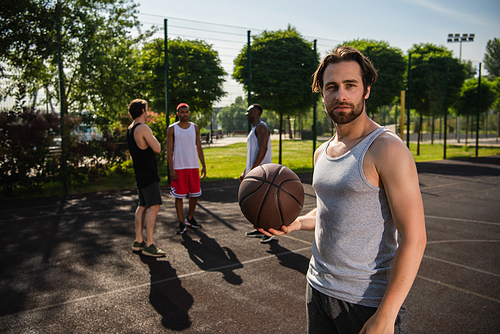Young man holding basketball ball near blurred interracial friends on playground