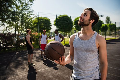 Sportsman with basketball ball looking away near blurred interracial friends on playground