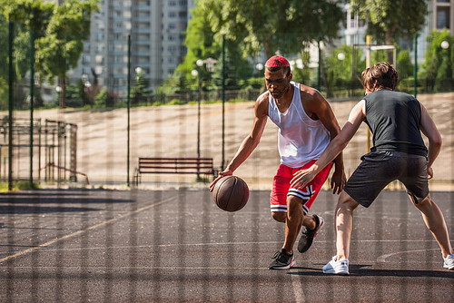 African american man playing streetball near friend and blurred fence
