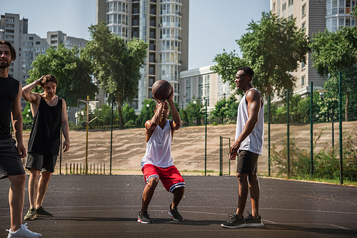 African american sportsman training with basketball ball near friends on playground on urban street
