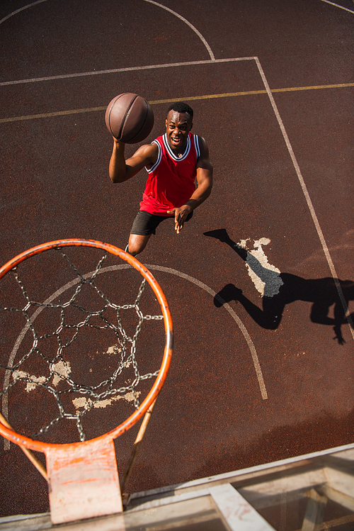 High angle view of excited african american sportsman jumping with basketball ball near hoop