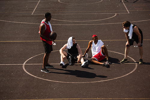 Smiling multiethnic sportsmen with basketball ball and water bottles talking on playground