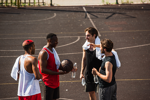 Cheerful african american man holding basketball ball and water near friends with towels on playground