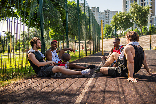 Cheerful interracial sportsmen with towels resting on basketball playground