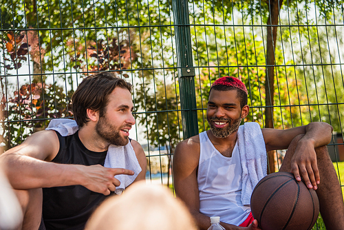 Young sportsman pointing with finger near african american friend with basketball outdoors