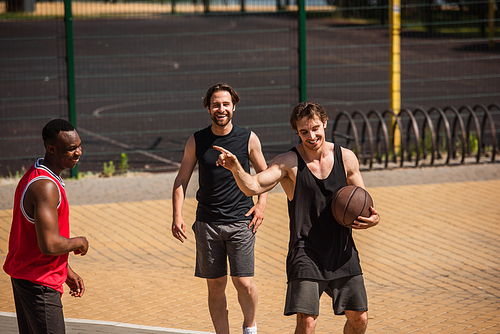 Smiling sportsman with basketball ball pointing with finger at african american friend outdoors