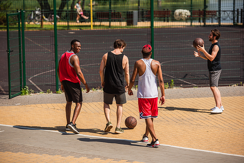 Cheerful multiethnic friends with basketball balls walking outdoors near playground