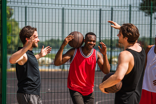 Positive multiethnic friends in sportswear holding basketball balls near fence outdoors