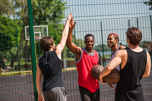 Smiling interracial sportsmen with basketball ball giving high five near fence outdoors