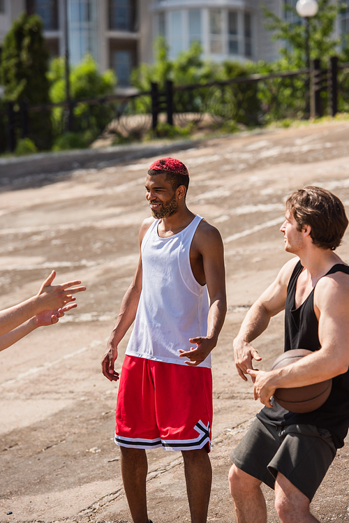 Smiling interracial men with basketball ball talking outdoors at daytime