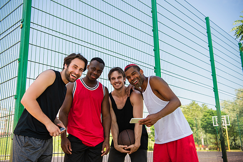 Cheerful multiethnic sportsmen with basketball ball and smartphones  outdoors