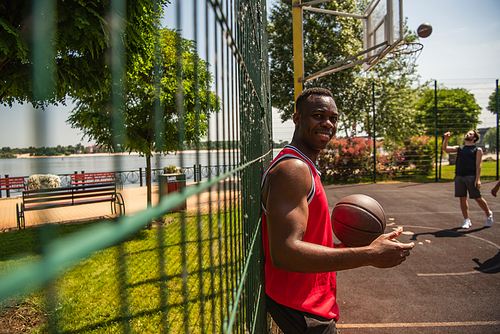 Young african american sportsman holding basketball ball near blurred fence on playground