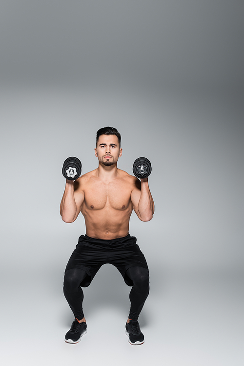 full length of strong sportsman doing squat with dumbbells on grey