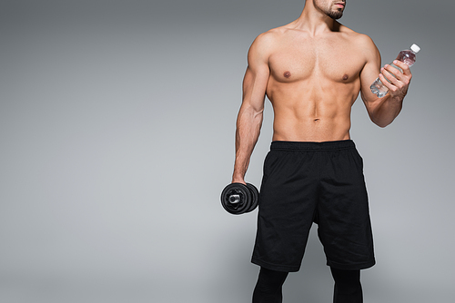 cropped view of shirtless sportsman holding dumbbell and bottle with water on grey