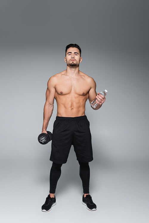 full length of shirtless sportsman holding dumbbell and bottle with water on grey