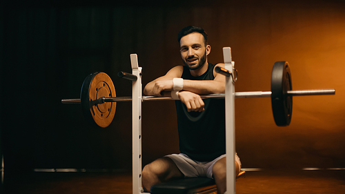 Smiling sportsman  near barbell on stand on dark background