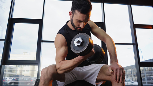 Low angle view of muscular sportsman training with dumbbell on tire in gym