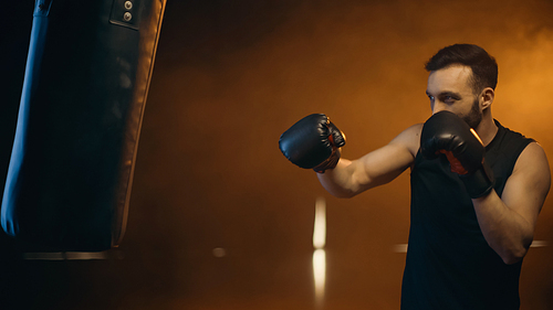 Sportsman in boxing gloves training with punch bag on dark background