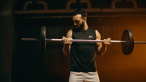 Muscular sportsman training with barbell on dark background