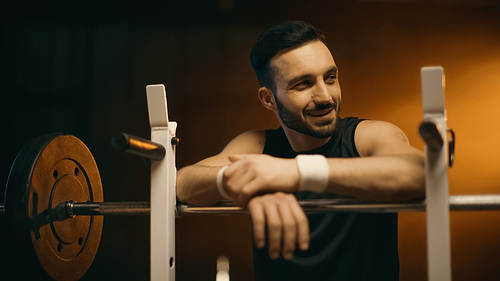 Smiling sportsman looking away near barbell on dark background