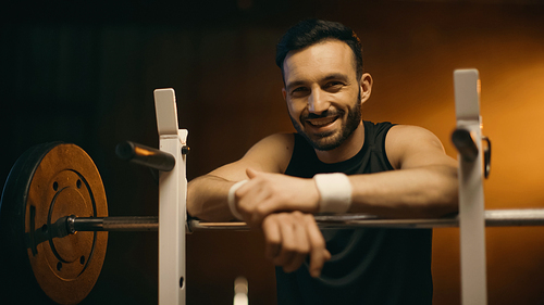 Sportsman smiling at camera near barbell on stand on dark background