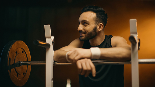 Smiling bearded sportsman standing near barbell on stand on dark background