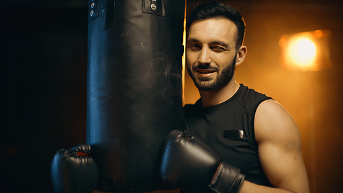 Smiling sportsman in boxing gloves winking near punch bag on dark background