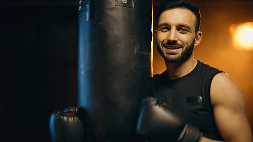 Positive sportsman in boxing gloves  near punch bag on dark background