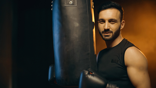 Muscular sportsman in boxing gloves  near punch bag on dark background