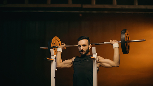 Muscular sportsman working out with barbell on dark background