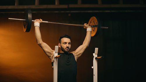 Muscular sportsman training with barbell near stand on dark background