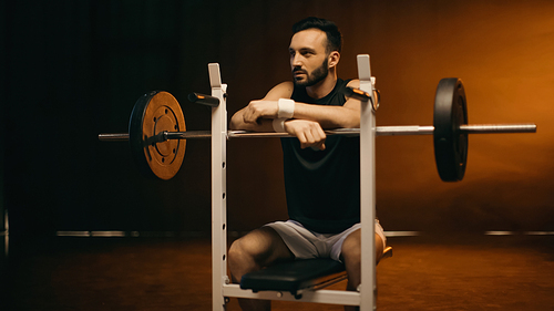 Bearded sportsman sitting on flat bench near barbell on dark background