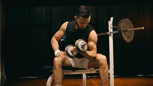 Muscular sportsman training with dumbbell on flat bench on dark background