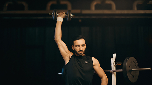 Athletic sportsman working out with dumbbell near barbell on dark background