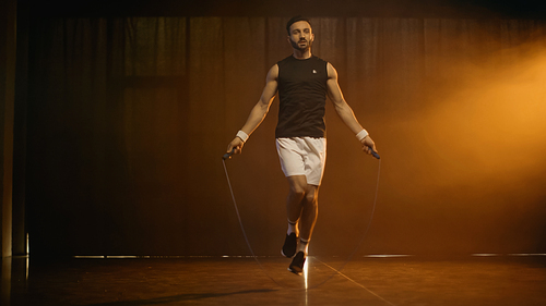 Athletic sportsman training with jump rope on dark background