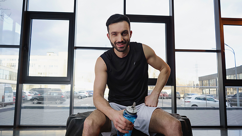 Smiling sportsman holding sports bottle while sitting on tire in gym