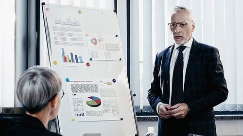 Mature businessman standing near blurred colleague and charts on flip chart in office