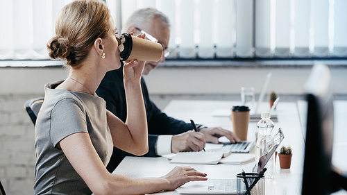 Businesswoman drinking coffee to go near laptop and blurred colleague in office