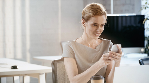 Smiling businesswoman using smartphone near blurred laptop and coffee to go in office