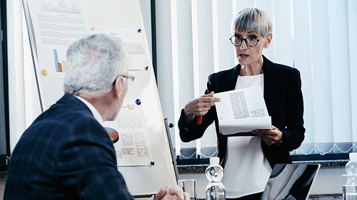 Mature businesswoman pointing at paper near colleague and flip chart in office