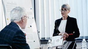 Middle aged businesswoman holding clipboard near colleague and flip chart in office