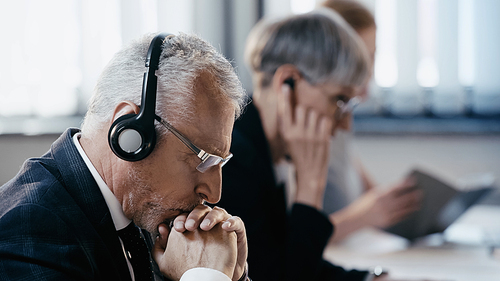 Side view of pensive businessman in headset sitting near blurred colleagues in office