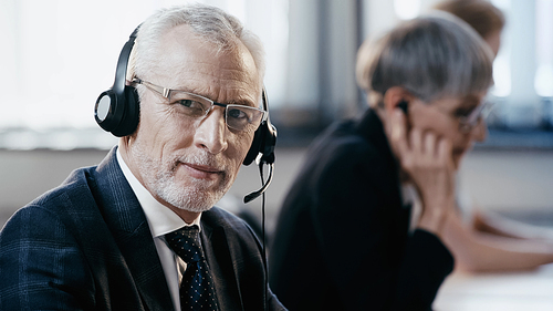 Mature businessman in headset  near blurred colleagues in office