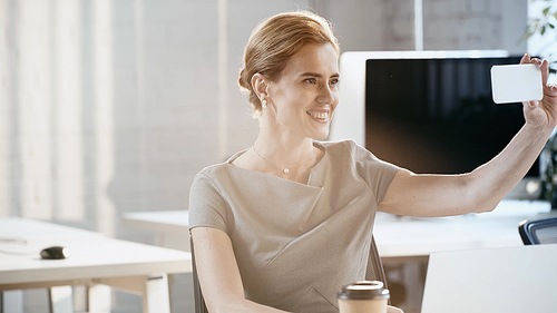 Positive businesswoman taking selfie on smartphone near laptop in office
