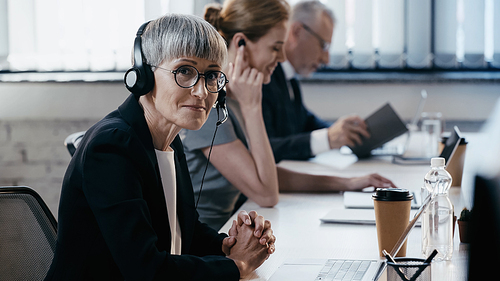 Mature businesswoman in headset  near laptop and coffee to go in office