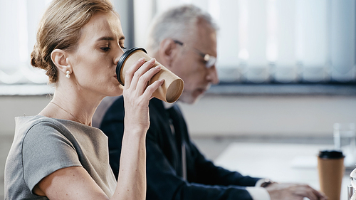 Businesswoman drinking coffee to go near blurred colleague working in office
